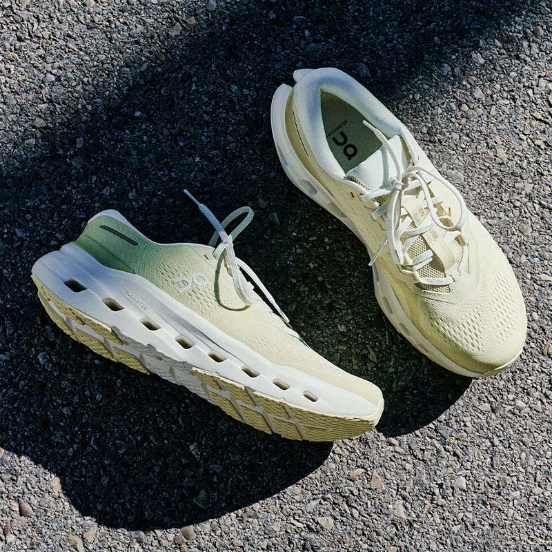 A pair of On Cloudrunner 3 running shoes in a pale yellow-green and off-white colorway, photographed from a high angle on a textured asphalt surface with strong natural shadows.
