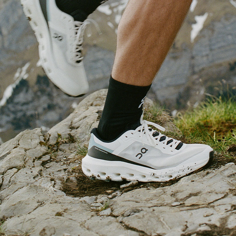 A close-up of a runner wearing On Cloudvista 3 trail running shoes with black socks, traversing a rocky outdoor landscape.