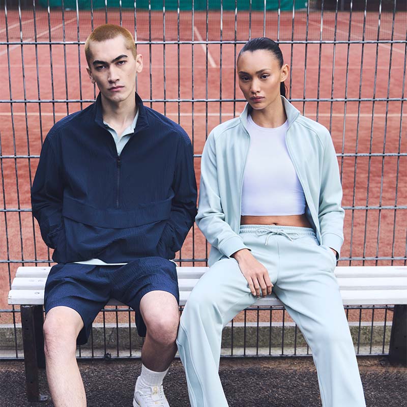 Two people sitting on a metal bench in front of a metal fence wearing On Court tennis clothing with a blurred clay tennis court background