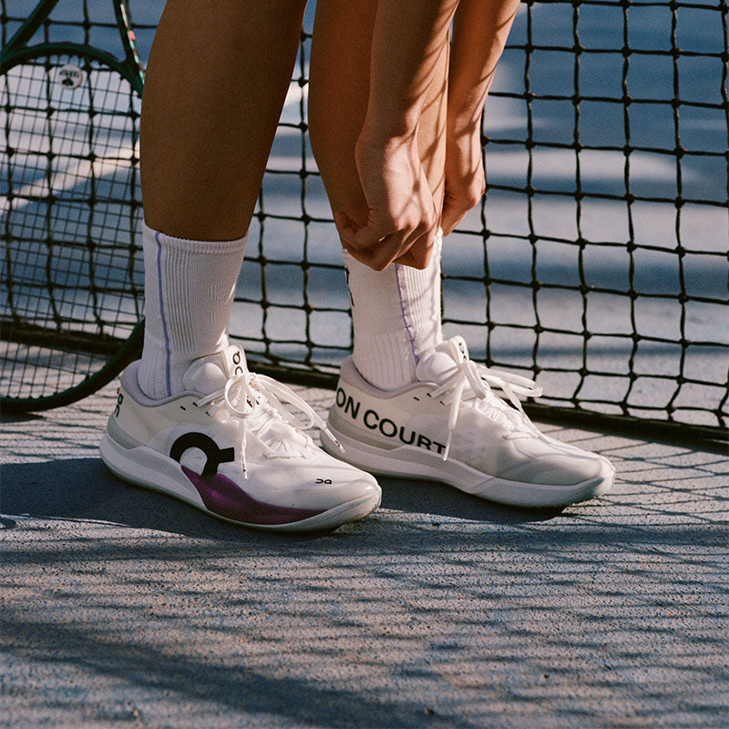 Closeup of a person wearing white On The Roger Pro 3 tennis shoes with purple accents and white crew socks on a blue gray court with a net in the background.