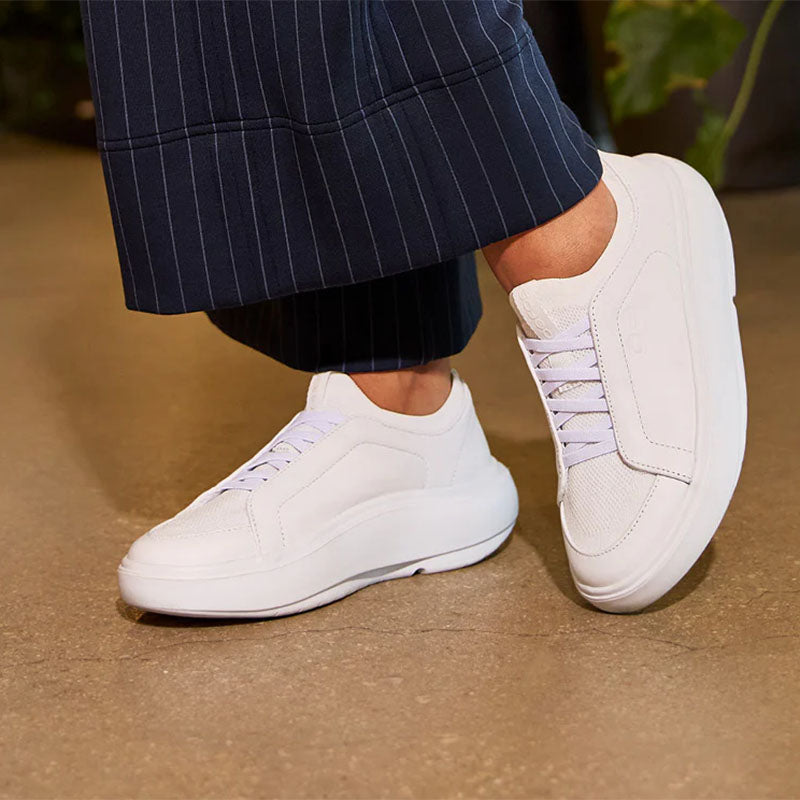 the feet of a person in dark blue pinstripe pants standing on a sand colored floor in white OOFOS Club+ walking shoes