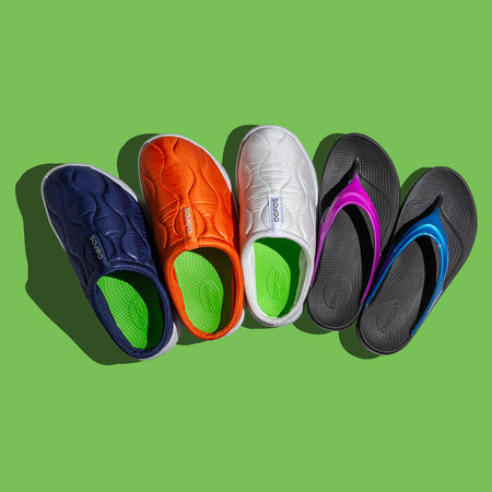 Colorful OOFOS recovery slip-on shoes and sandals arranged in a row on a green background