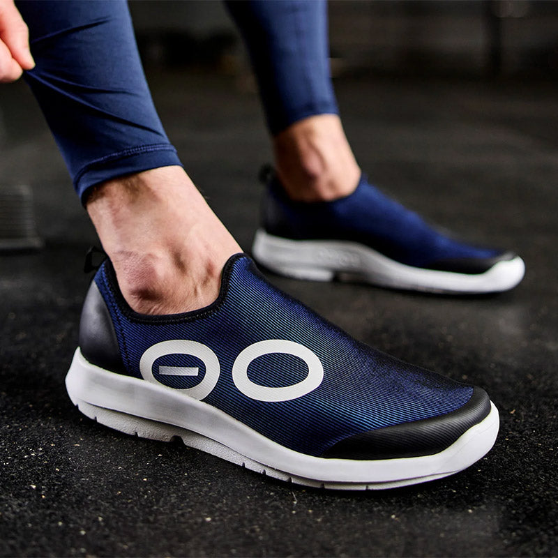 Closeup of a person wearing white/navy OOFOS OOmg Sport recovery shoes on a black gym floor.