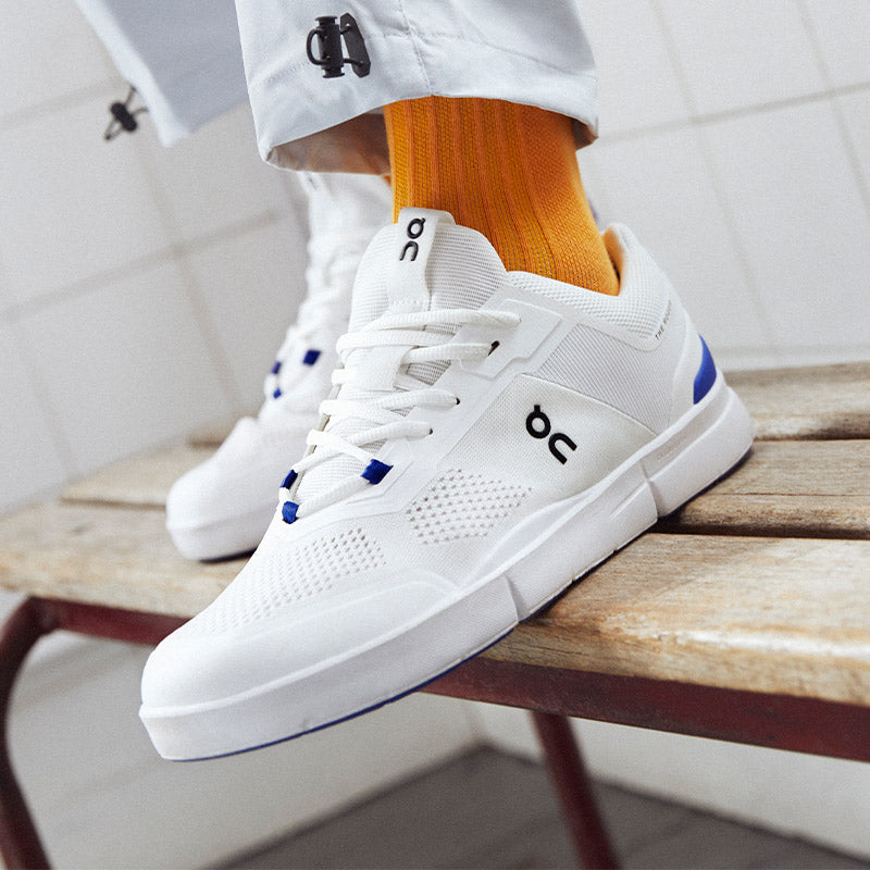 Lifestyle image: a person standing on a lockerroom bench in white and blue On The Roger Spin lifestyle shoes.
