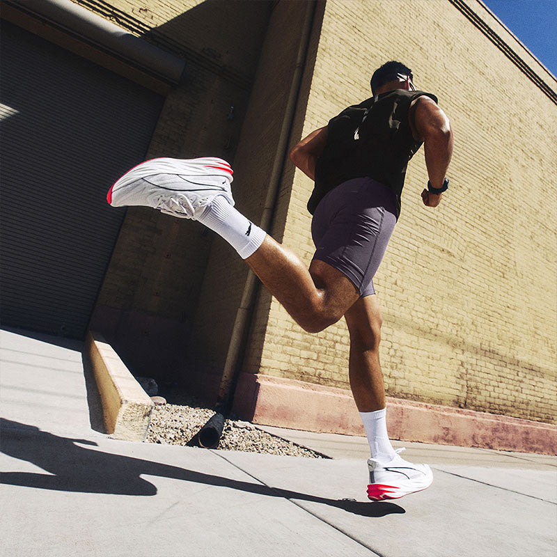 A runner on a sidewalk in front of a brick building wearing white Puma Deviate Pure NITRO running shoes with red and black accents