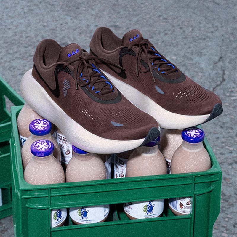 A pair of R.A.D UFO 'Choccy Milk' Unisex running shoes, displayed on top of a green crate containing chocolate milk bottles, set against a gray asphalt background.