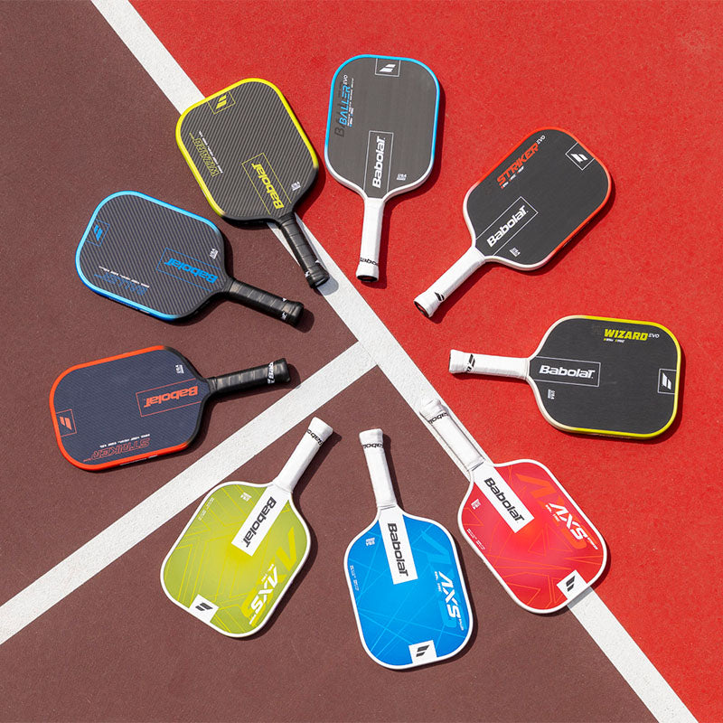 A collection of Babolat pickleball paddles in various colors and models arranged in a circular pattern on a red pickleball court background.