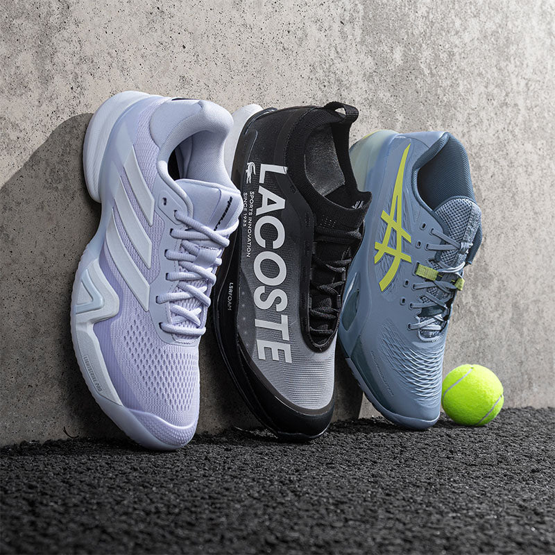 Three tennis shoes leaning against a textured concrete wall on a dark surface: a light purple adidas shoe on the left, a black and gray Lacoste shoe in the center, and a blue ASICS shoe on the right with a yellow tennis ball beside it.