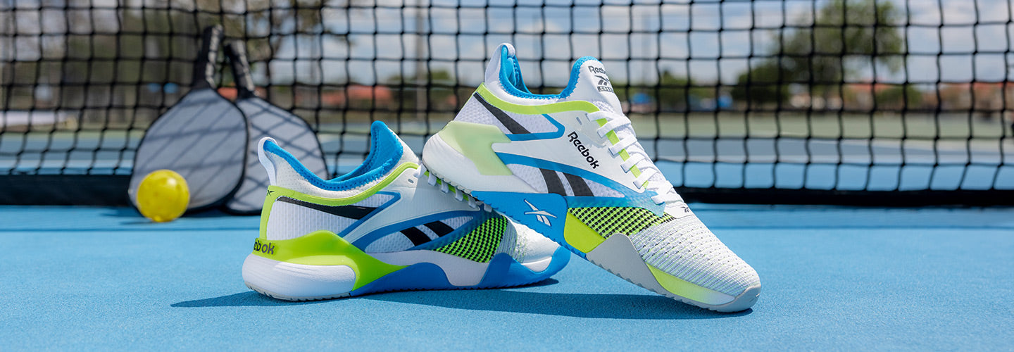 Reebok Nano Court men's shoes sitting on a pickleball court in front of a pickleball net with two pickleball paddles and yellow pickleball behind them.