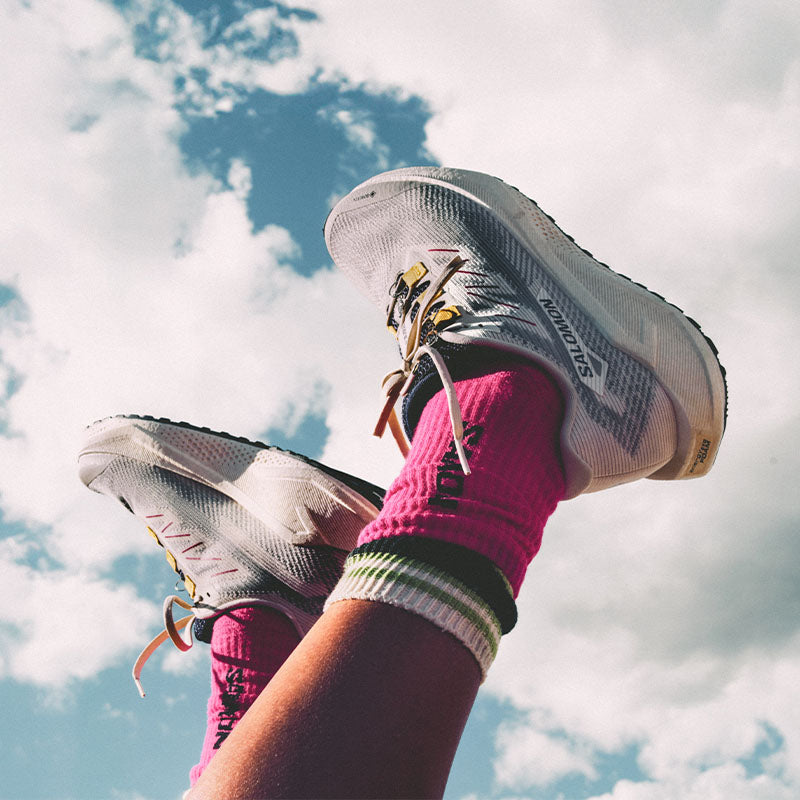 A person their feet in the air, wearing white Salon Aero Glide GRVL GTX running shoes with colorful socks against a cloudy sky