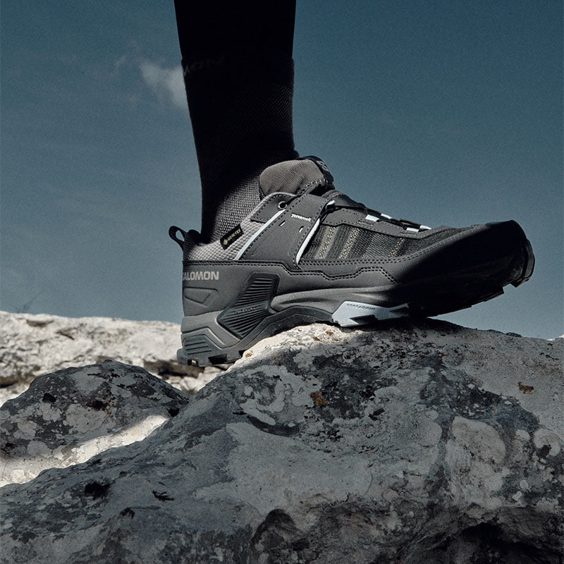 a hiker standing on a rocky surface wearing Salomon X Ultra 5 GTX hiking shoes with a blue sky in the background