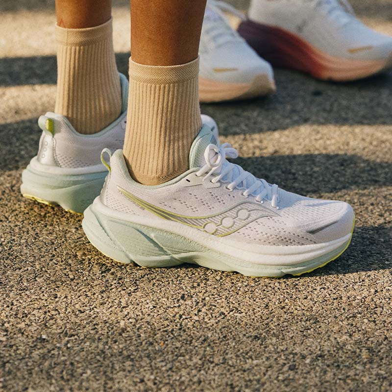 Person wearing white Saucony Hurricane 25 running shoes with green and yellow accents, standing on paved surface next to another person in the background