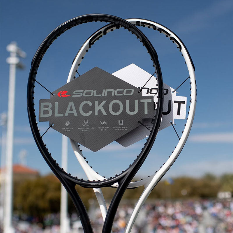 Two unstrung Solinco Blackout and Whiteout v1 tennis racquets with a blurred sky and spectator seating in the background