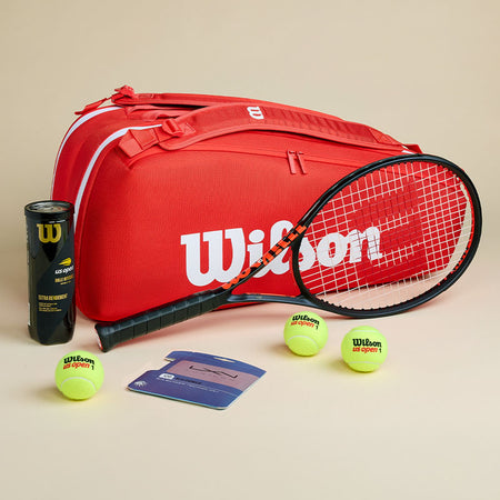 Red Wilson tennis bag with black Clash racquet and a Luxilon string package on a light background.