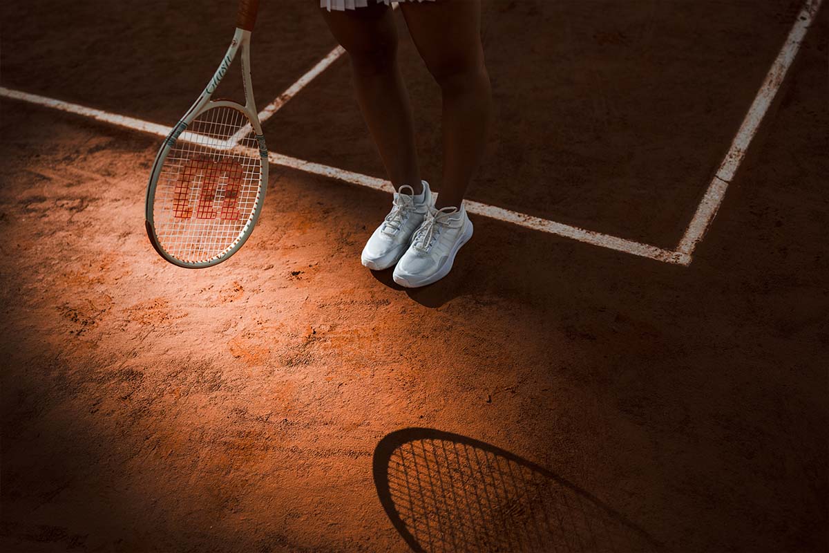 Woman holding a Wilson Clash Roland Garros tennis racquet and wearing white Wilson Intrigue tennis shoes on a clay court