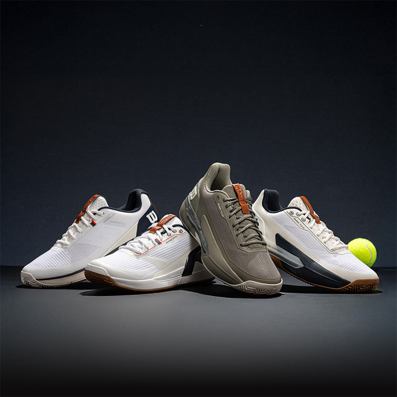 Four Wilson Rush tennis shoes arranged on a dark studio surface against a black background, with a yellow tennis ball near the right shoe.